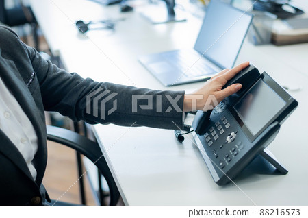 Closeup female hand on landline phone in office. Faceless woman in a suit works as a receptionist answering the phone to customer calls. 88216573