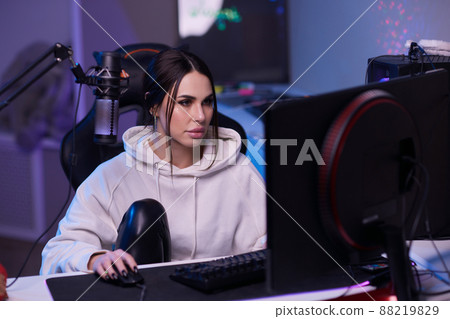 Happy woman putting on headphones and playing computer game. Female gamer in glasses sitting and looking at computer monitor. 88219829
