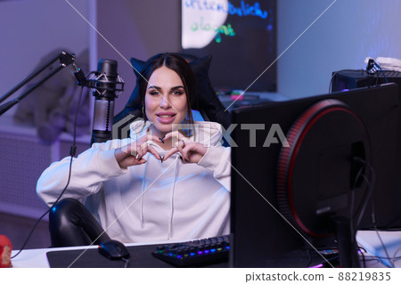 Happy woman putting on headphones and playing computer game. Female gamer in glasses sitting and looking at computer monitor. Happy woman putting on headphones and playing computer game. Female gamer in glasses sitting and looking at computer monitor. 88219835