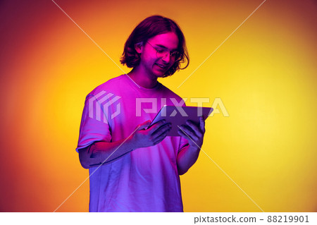 Portrait of young man with long hair using digital gadget isolated on orange background in purple neon light. Concept of emotions, beauty, fashion Portrait of young man with long hair using digital gadget isolated on orange background in purple neon light. Concept of emotions, beauty, fashion 88219901