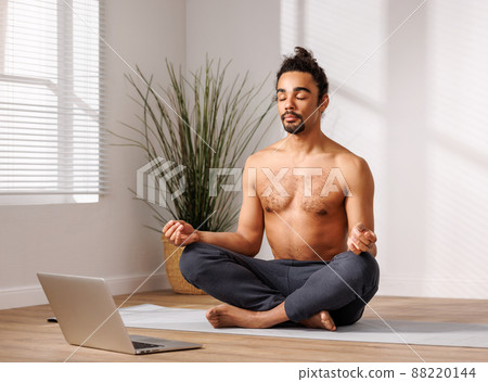 Relaxed black man meditating in lotus pose in front of laptop at home Relaxed black man meditating in lotus pose in front of laptop at home 88220144