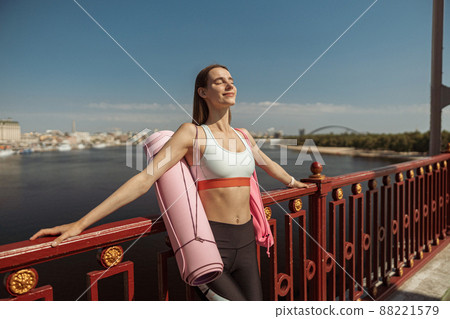 Happy peaceful woman with mat and backpack leans onto handrail on city embankment Happy peaceful woman with mat and backpack leans onto handrail on city embankment 88221579