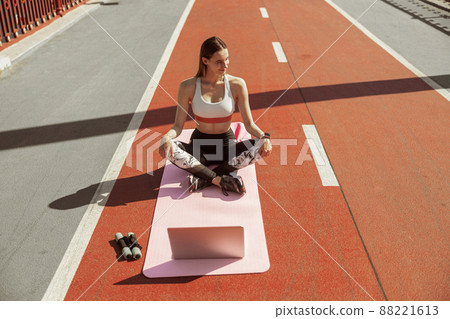 Sportswoman with laptop and dumbbells sits on sportsground on sunny day 88221613