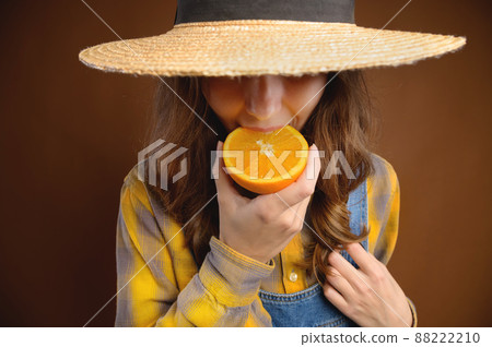 Studio portrait of an attractive caucasian young woman in a hat and denim overalls holds a cut orange fruit in her hands and shows her tongue trying to taste it Studio portrait of an attractive caucasian young woman in a hat and denim overalls holds a cut orange fruit in her hands and shows her tongue trying to taste it 88222210