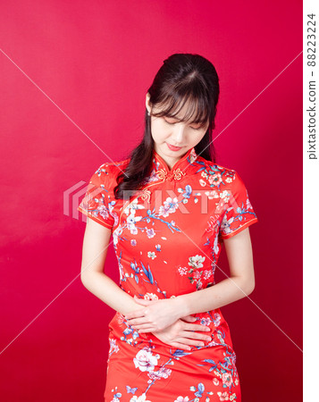 Portrait of a young asian woman in traditional cheongsam qipao dress having a stomach ache isolated over red background. Portrait of a young asian woman in traditional cheongsam qipao dress having a stomach ache isolated over red background. 88223224