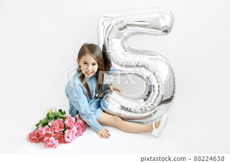 A long-haired brunette girl is sitting with a bouquet of flowers and a large balloon in the shape of the number five. A cute cute child is celebrating his first anniversary - the fifth anniversary. A long-haired brunette girl is sitting with a bouquet of flowers and a large balloon in the shape of the number five. A cute cute child is celebrating his first anniversary - the fifth anniversary. 88224638