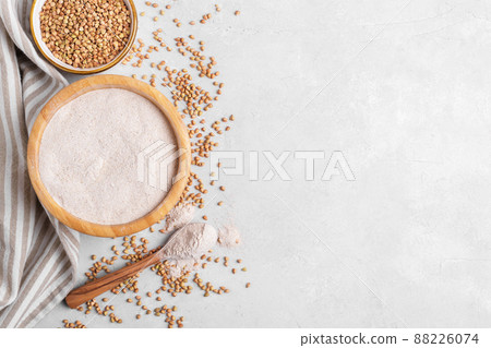 Buckwheat flour in a wooden bowl with wooden spoon and raw green buckwheat grain in a bowl on grey stone background Buckwheat flour in a wooden bowl with wooden spoon and raw green buckwheat grain in a bowl on grey stone background 88226074