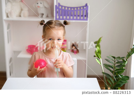 A two-year-old child breathes into an inhalation mask. Inhalations for asthma, bronchitis and pneumonia. Treatment of children's diseases of the upper respiratory tract. A toy in the shape of a virus A two-year-old child breathes into an inhalation mask. Inhalations for asthma, bronchitis and pneumonia. Treatment of children's diseases of the upper respiratory tract. A toy in the shape of a virus 88226313