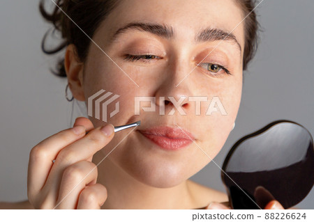 A young woman looks in the mirror and plucks her hair over her upper lip with tweezers. The concept of getting rid of unwanted facial hair. Close up 88226624