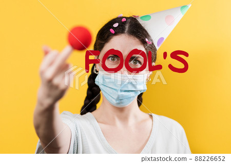 April's fool day. Angry woman in a paper cap and protective mask, showing the middle finger with a red pompom. Yellow background. Coronavirus new normal concept April's fool day. Angry woman in a paper cap and protective mask, showing the middle finger with a red pompom. Yellow background. Coronavirus new normal concept 88226652