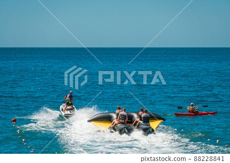 Happy people are going to swim on an air mattress behind a motor boat. Tourists ride the inflatable watercraft boat. Sea attraction. Power boat skating people on a large Inflatable raft. 88228841