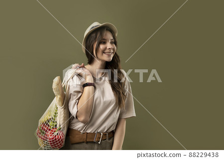 Smiling young woman in light summer clothes with a mesh eco bag full of vegetables, greens watching in a camera on a green studio background. Sustainable lifestyle. Eco friendly concept. Zero waste. Smiling young woman in light summer clothes with a mesh eco bag full of vegetables, greens watching in a camera on a green studio background. Sustainable lifestyle. Eco friendly concept. Zero waste. 88229438