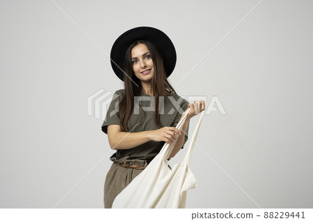 Happy smiling brunette woman in black hat with white cotton eco bag on her shoulder. Girl holding textile grocery bag with vegetables. Zero waste concept. 88229441
