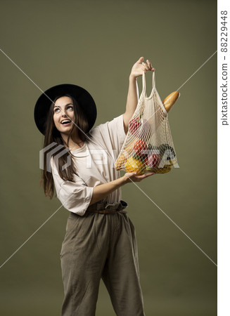 Eco friendly shopping. Smiling young woman holding string reusable mesh bag with organic fruits and vegetables. Positive female standing on green studio background. Eco friendly shopping. Smiling young woman holding string reusable mesh bag with organic fruits and vegetables. Positive female standing on green studio background. 88229448