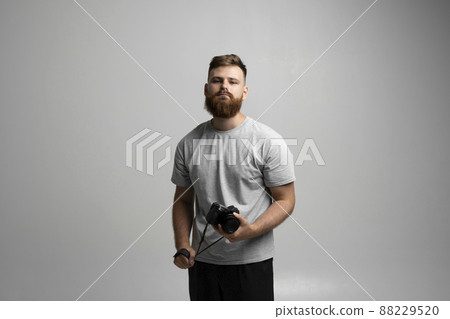 Portrait of bearded professional photographer with dslr camera looks straight into the camera isolated on gray background. 88229520