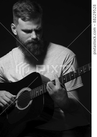 Bearded brutal guitarist plays an acoustic guitar in a black room. The concept of music recording, rehearsal or live performance. 88229528