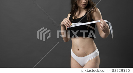 Cropped image beautiful young brunette woman 20s in black and white underwear standing posing measuring breast with measure tape isolated on black wall background. 88229550