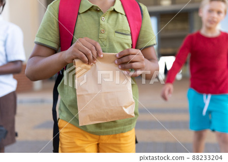 Midsection of biracial elementary schoolboy with packed lunch standing in school campus 88233204