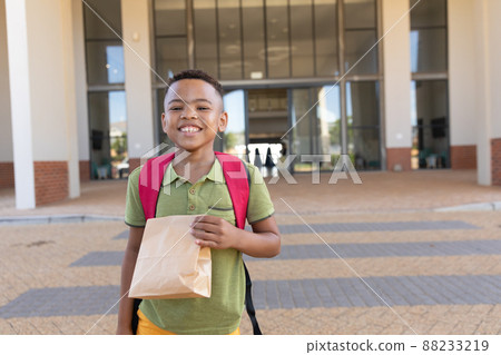Portrait of smiling biracial elementary schoolboy with packed lunch standing in school campus 88233219
