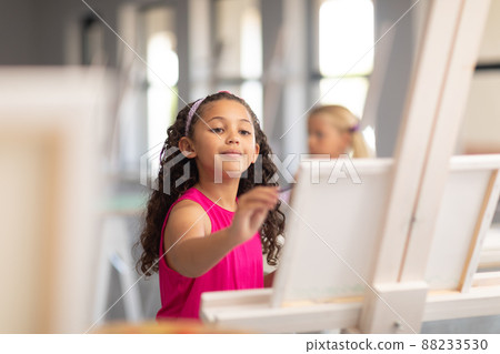 Cute biracial elementary schoolgirl painting on easel during drawing class in school 88233530