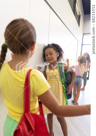 Multiracial elementary schoolgirls talking while standing by lockers in school corridor 88233552