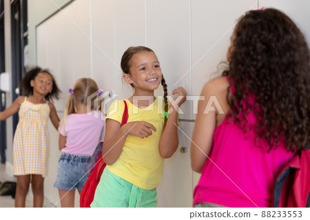 Multiracial elementary schoolgirls talking while standing by lockers in school corridor Multiracial elementary schoolgirls talking while standing by lockers in school corridor 88233553