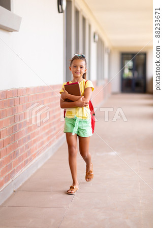Full length portrait of caucasian elementary schoolgirl with books and backpack walking in corridor Full length portrait of caucasian elementary schoolgirl with books and backpack walking in corridor 88233671