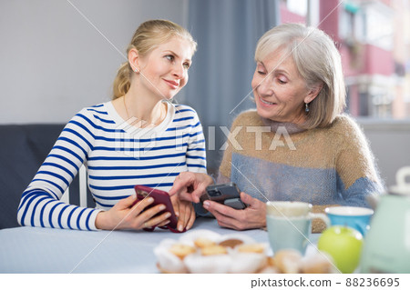 Mature mother and daughter together playing on their smartphones while sitting at table at home Mature mother and daughter together playing on their smartphones while sitting at table at home 88236695
