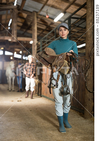 Woman jockey with saddle and bridle in her hands in stable 88237189