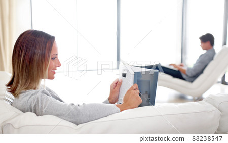 Catching up on the latest fashions. A young woman reading a magazine while her boyfriend browses a laptop in the background. Catching up on the latest fashions. A young woman reading a magazine while her boyfriend browses a laptop in the background. 88238547