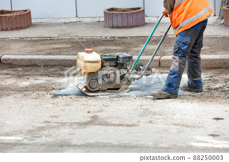 A road worker is patching up a pothole in an old road using an old petrol plate compactor. 88250003