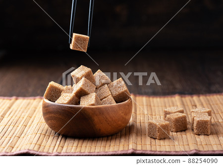 Cube of cane sugar between chopsticks over a bowl of sugar. 88254090