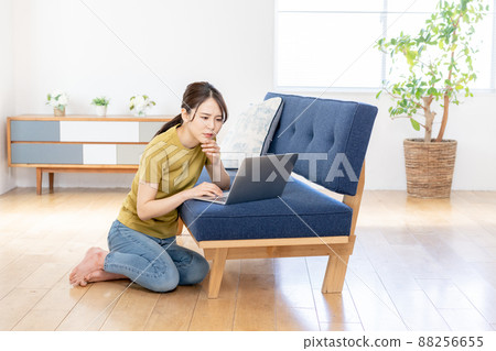 A troubled-faced young woman sitting on the floor of the living room and working on a laptop 88256655