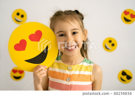 World emoji day. Anthropomorphic smile Face. Birthday Party. Emotions. A little girl, smiling with all her teeth, holds a cardboard love emoji in her hands. World emoji day. Anthropomorphic smile Face. Birthday Party. Emotions. A little girl, smiling with all her teeth, holds a cardboard love emoji in her hands. 88256656