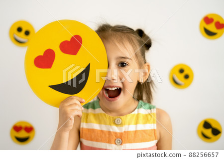 A little girl, smiling with her mouth open, holds in her hands a cardboard love smile with hearts instead of eyes, covering part of her face with them. World emoji day. Anthropomorphic smile Face. 88256657