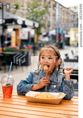 A little stylish girl in a denim jacket eats harmful fast food outdoors on the terrace of a cafe. The child is happy and enjoys eating fried chicken, washed down with a refreshing summer lemonade 88256998