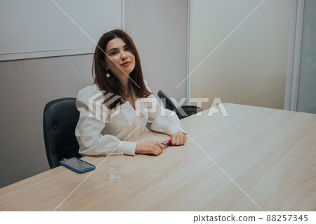Young brunette woman in a white blouse sitting at the table in the office, holding a marker in her hands and looking at the camera. Business Education concept. Young brunette woman in a white blouse sitting at the table in the office, holding a marker in her hands and looking at the camera. Business Education concept. 88257345
