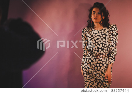 Beautiful brunette fashion model in polka dot dress posing for the photographer on a pink background. Shadow and glare on the wall Beautiful brunette fashion model in polka dot dress posing for the photographer on a pink background. Shadow and glare on the wall 88257444