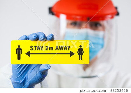 Medical worker in personal protective equipment holding yellow "STAY 2m AWAY" sign Medical worker in personal protective equipment holding yellow "STAY 2m AWAY" sign 88258436