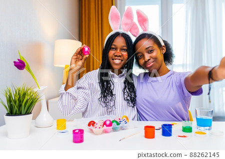 multiracial two women painting eggs with cute pink rabbit costume ears taking selfie photo on camera of smartphone in apartment 88262145