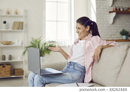 Happy young woman sitting on sofa and talking to someone on video call on laptop Happy young woman sitting on sofa and talking to someone on video call on laptop 88262889
