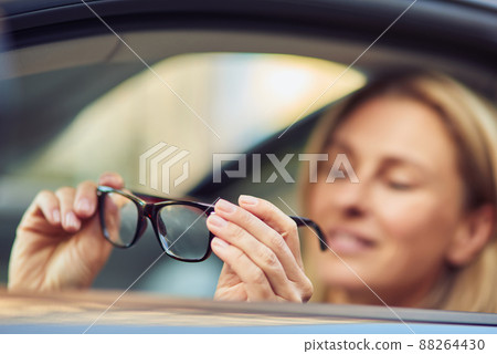 Portrait of a happy business woman holding eyeglasses and smiling at camera while sitting behind steering wheel of her modern car, selective focus Portrait of a happy business woman holding eyeglasses and smiling at camera while sitting behind steering wheel of her modern car, selective focus 88264430