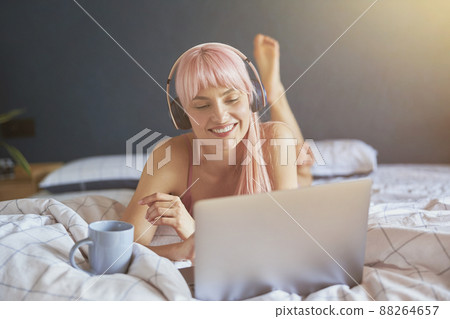 Positive woman with headphones and cup watches video via laptop on bed Positive woman with headphones and cup watches video via laptop on bed 88264657