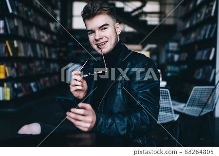 Positive young man with smartphone looking at camera in library Positive young man with smartphone looking at camera in library 88264805