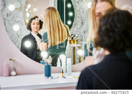 Young woman looking in the mirror and smiling while esthetician measuring eyebrow distance with brow ruler. Beauty specialist using measuring tool before eyebrow correction procedure in beauty salon. Young woman looking in the mirror and smiling while esthetician measuring eyebrow distance with brow ruler. Beauty specialist using measuring tool before eyebrow correction procedure in beauty salon. 88275925