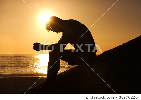 Contemplating the meaning of life. Silhouette of a man sitting by the ocean and contemplating while looking downwards. Contemplating the meaning of life. Silhouette of a man sitting by the ocean and contemplating while looking downwards. 88276226