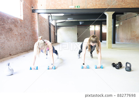 Two young attractive women doing push ups inside with barbells. Fitness woman working out in a cross Two young attractive women doing push ups inside with barbells. Fitness woman working out in a cross 88277698