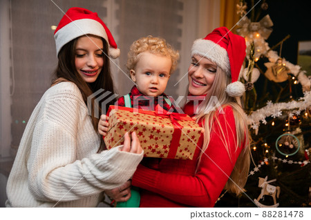 A mom and big sister give a gift to a two-year-old child at Christmas. A mom and big sister give a gift to a two-year-old child at Christmas. 88281478