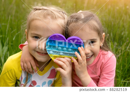 Two little girls play with a modern heart-shaped pop it toy. A fascinating sensory toy for the development of fine motor skills . Simple simple. Two girl friends have fun in nature. Two little girls play with a modern heart-shaped pop it toy. A fascinating sensory toy for the development of fine motor skills . Simple simple. Two girl friends have fun in nature. 88282519