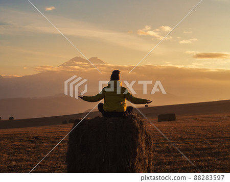 A woman sits on a haystack with her arms spread apart against the backdrop of Elbrus in the clouds at sunset. View from the back A woman sits on a haystack with her arms spread apart against the backdrop of Elbrus in the clouds at sunset. View from the back 88283597